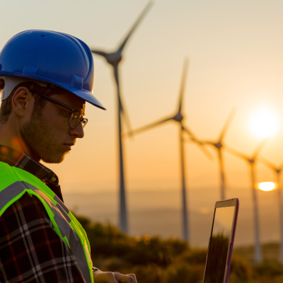 A focused male engineer in high visibility clothing inspecting renewable energy operations on a tablet at a wind farm during a vibrant sunset.